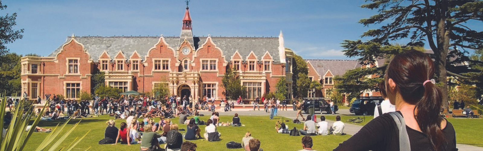 Hundreds of students are sitting on a green lawn in front of a red brick building with ornate points throughout. 